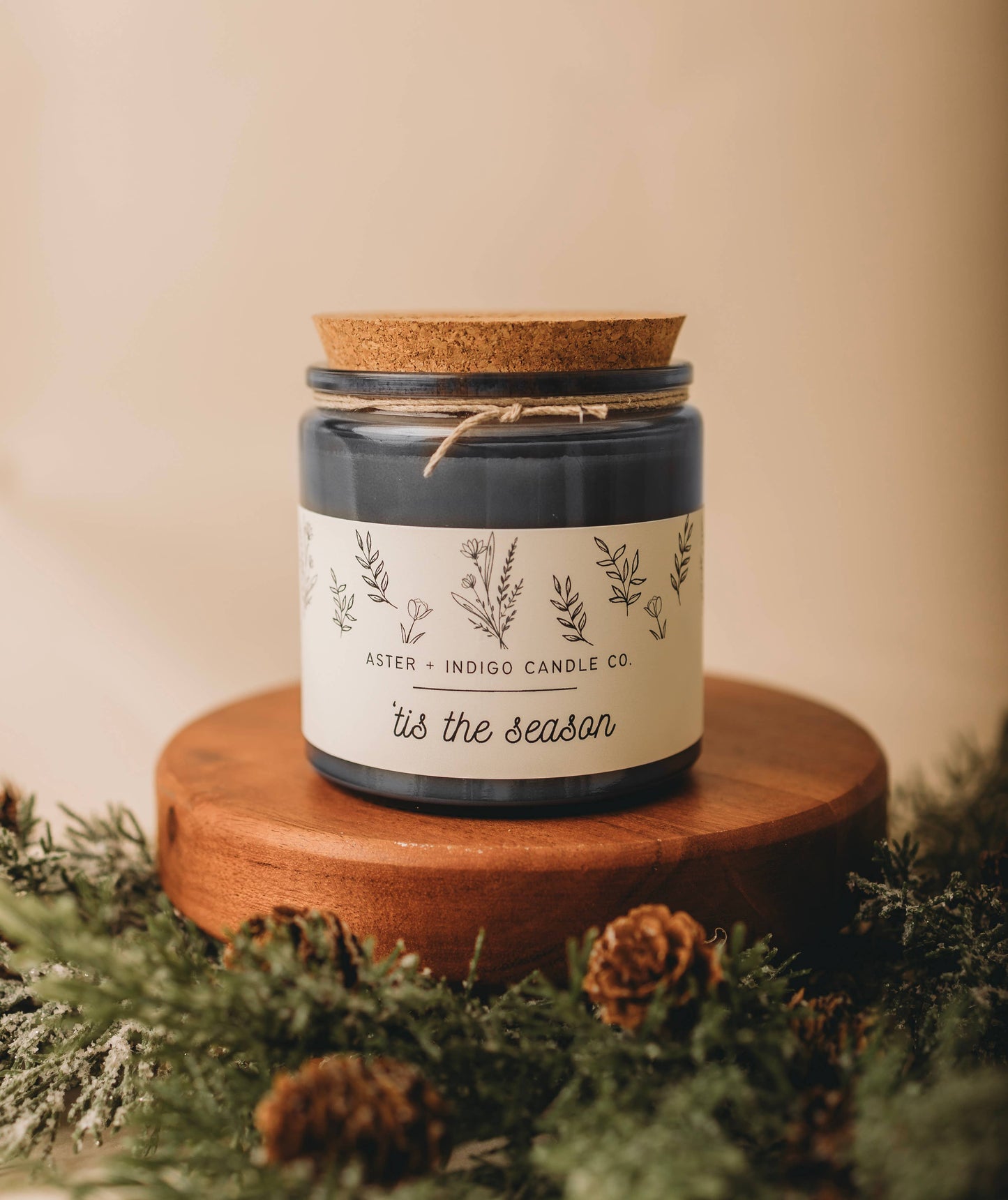 Candle jar with decorative label on a wooden stand surrounded by greenery