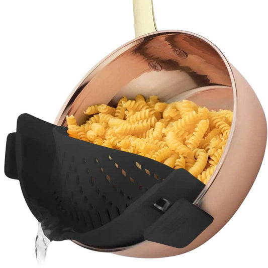 A copper-colored saucepan with a black silicone pasta strainer basket, shown with rotini pasta inside and water draining from the strainer against a white background.