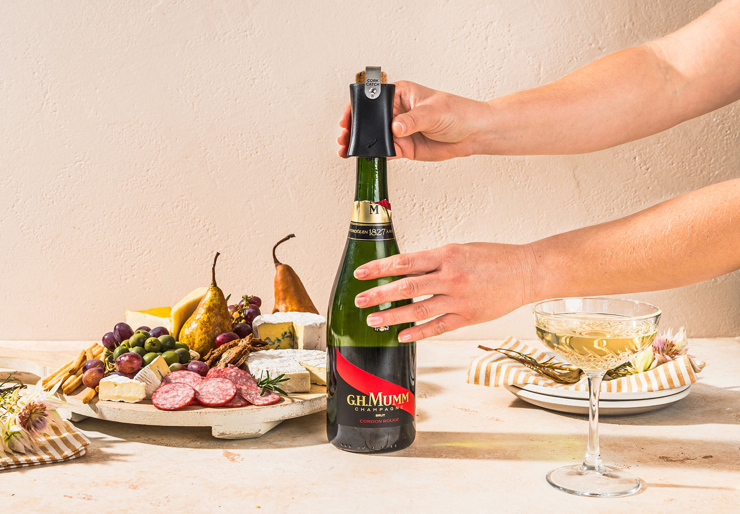Person opening a bottle of champagne using the Dreamfarm Champop with a charcuterie board and glass of champagne in the background.