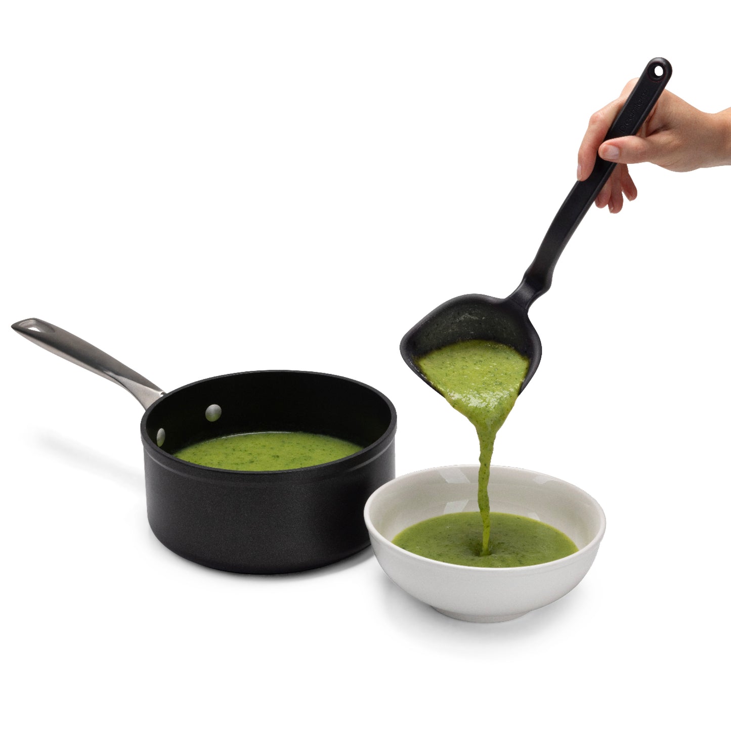 Green soup being poured from a ladle into a bowl with a black pan on a white background