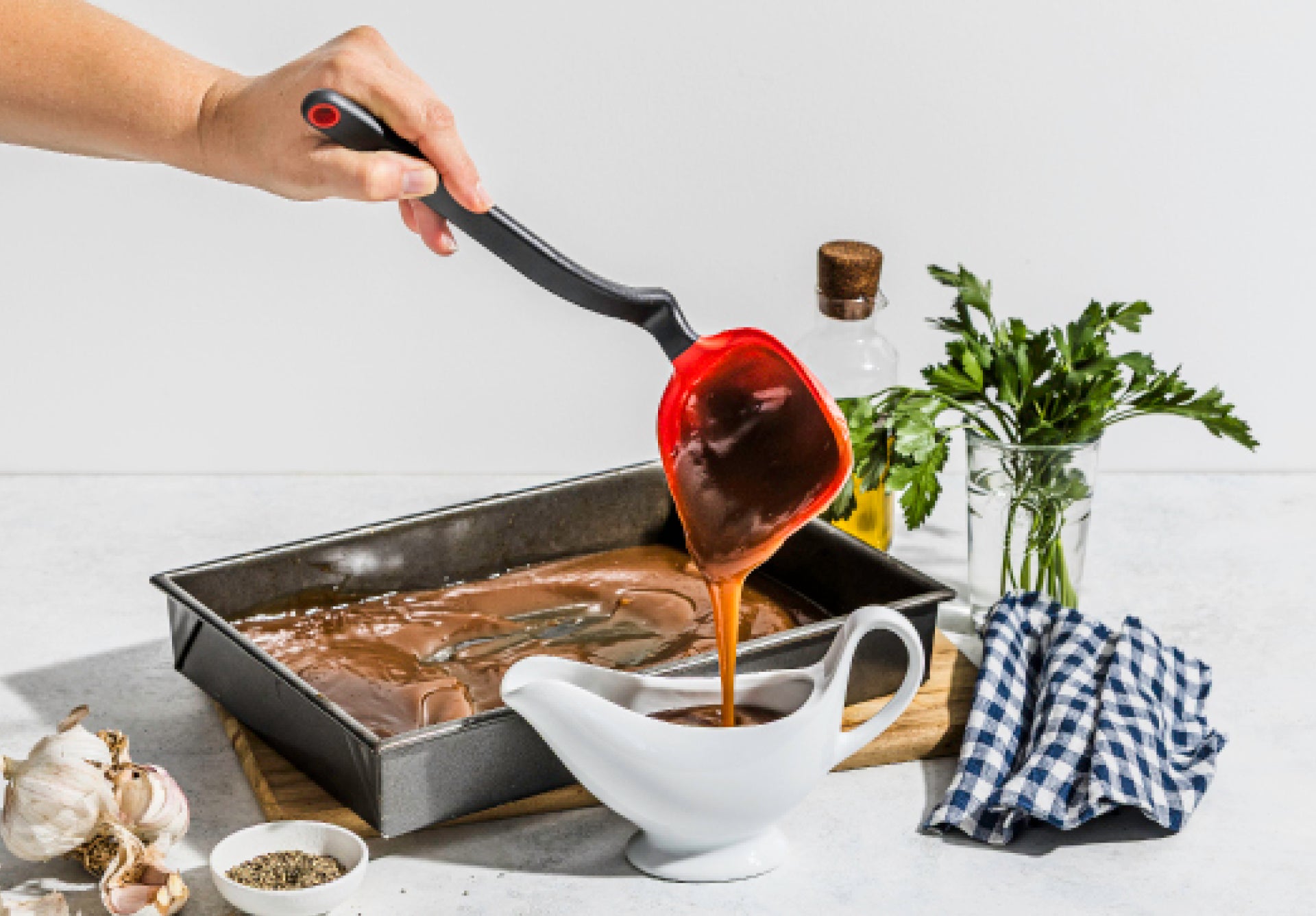 Person pouring red sauce from a ladle into a white gravy boat on a kitchen counter.