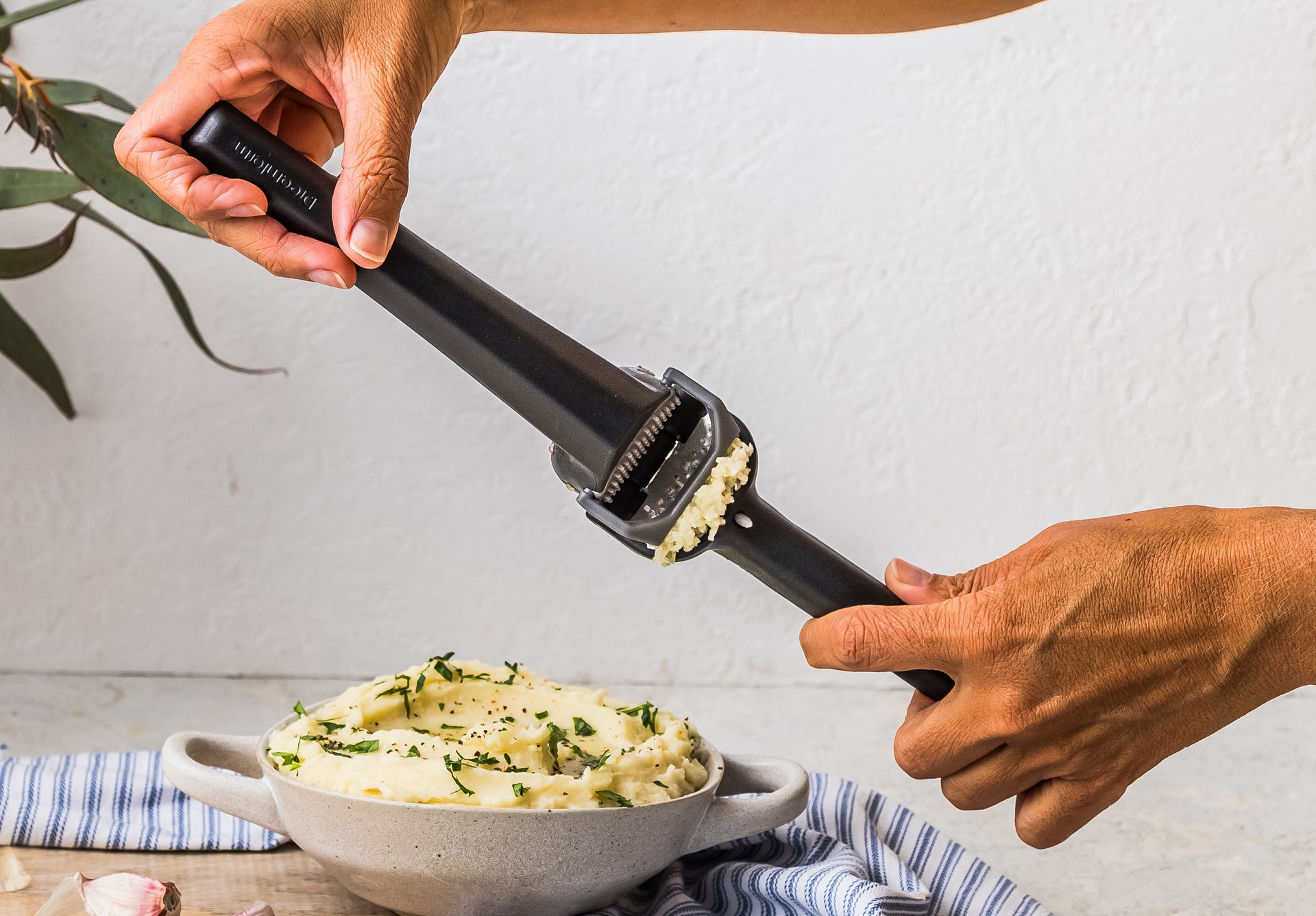Person using a garlic press to make minced garlic over a bowl of mashed potatoes.