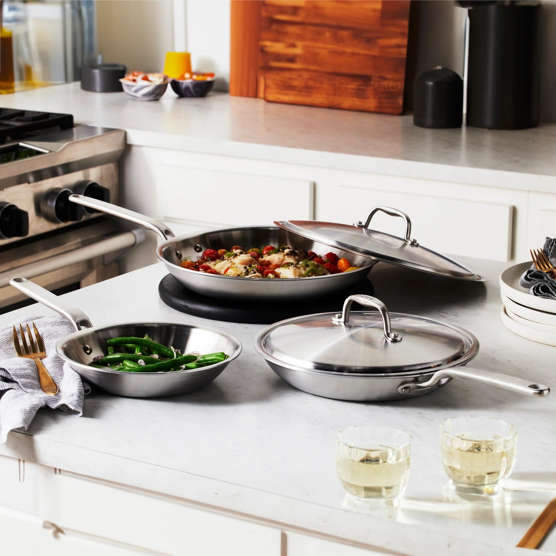 Set of stainless steel cookware on a kitchen counter with food and glasses.