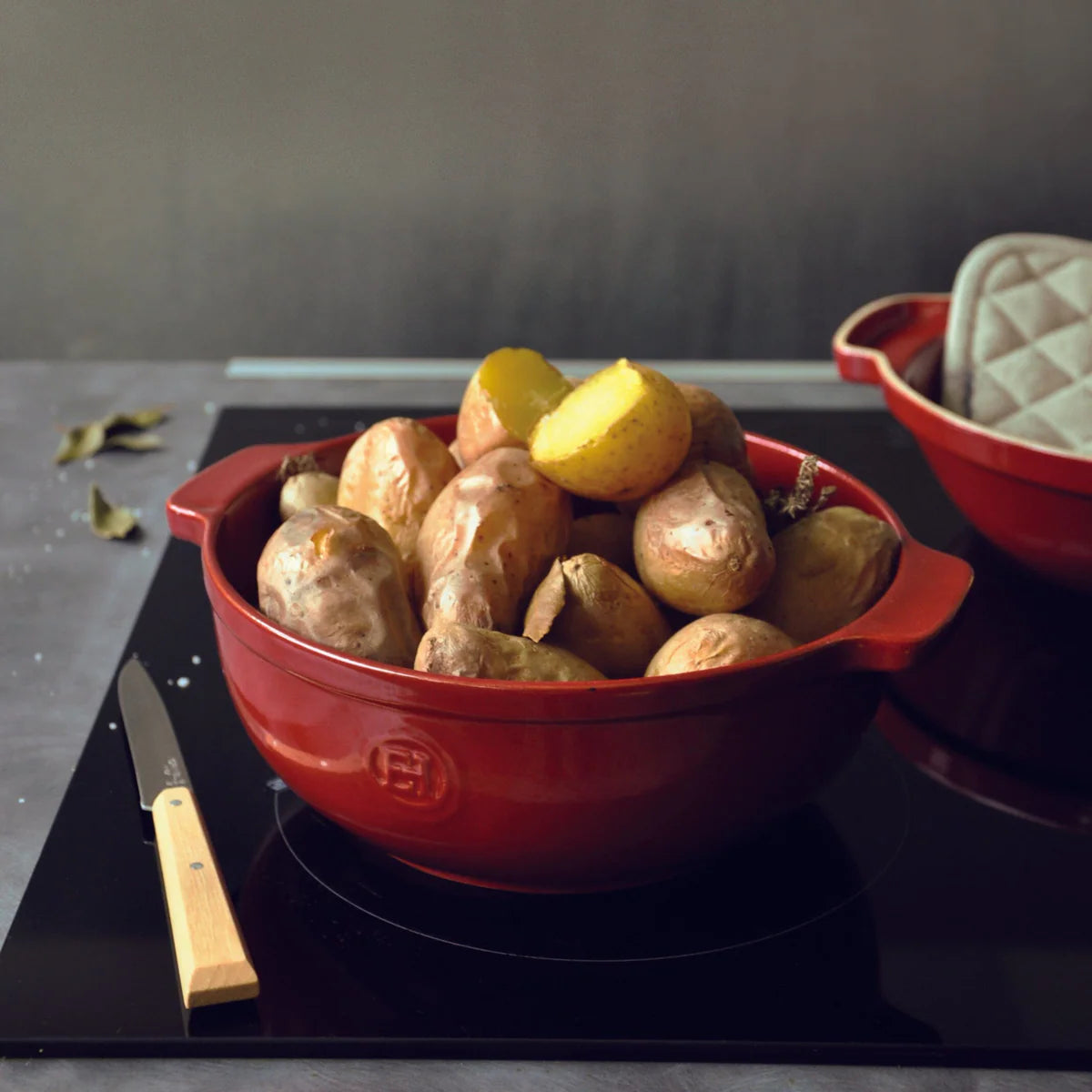 Red bowl filled with potatoes on a stove, with a knife and oven mitt in the background.