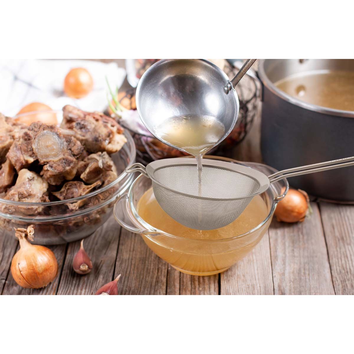 Bowl of broth with a ladle, onions, and bones on a wooden surface