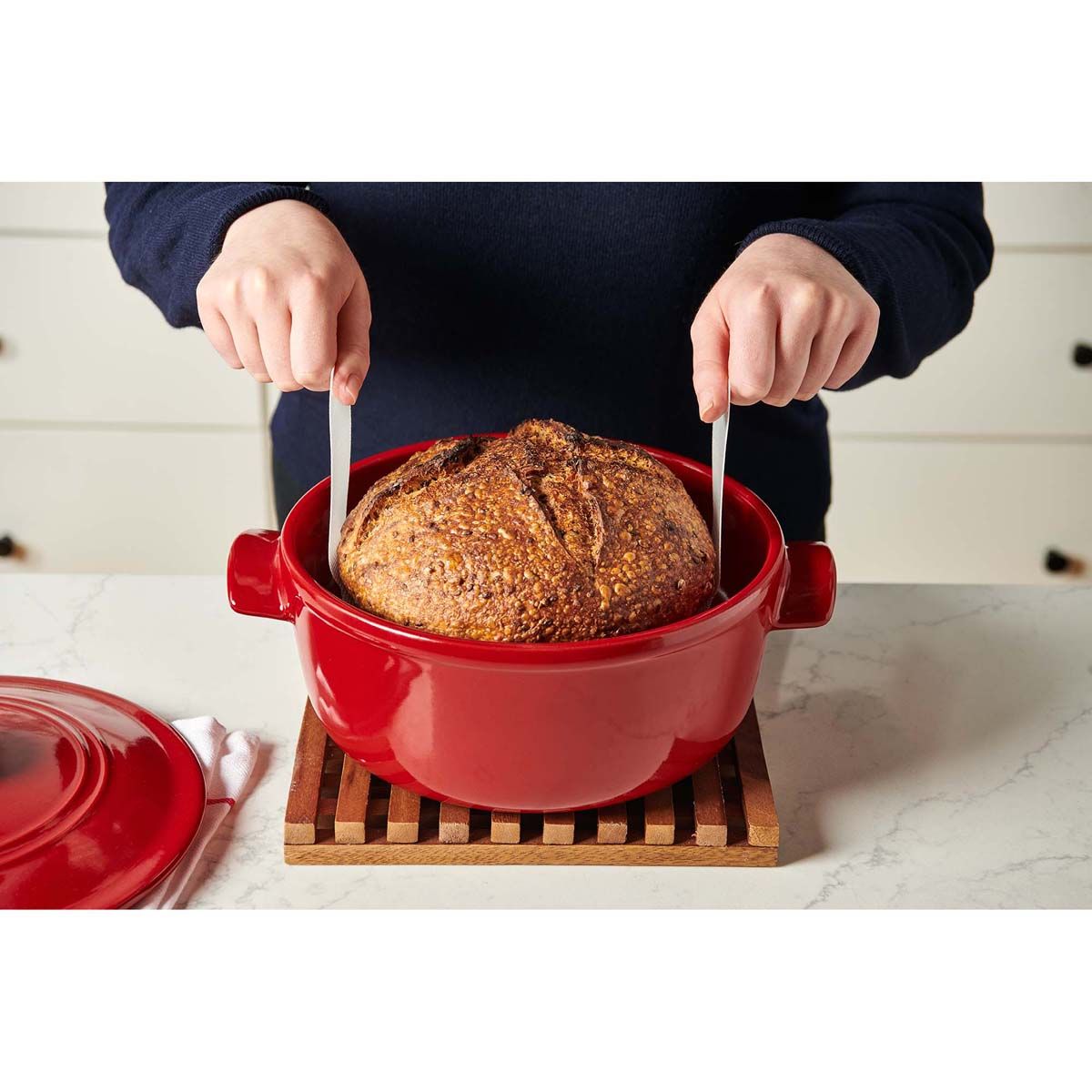Person lifting a loaf of bread using the loaf lifter in a red ceramic dish on a marble countertop.