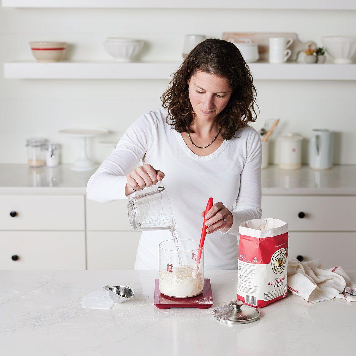Woman in a kitchen pouring flour into a sourdough crock with a red spatula and a bag of flour on a white countertop.