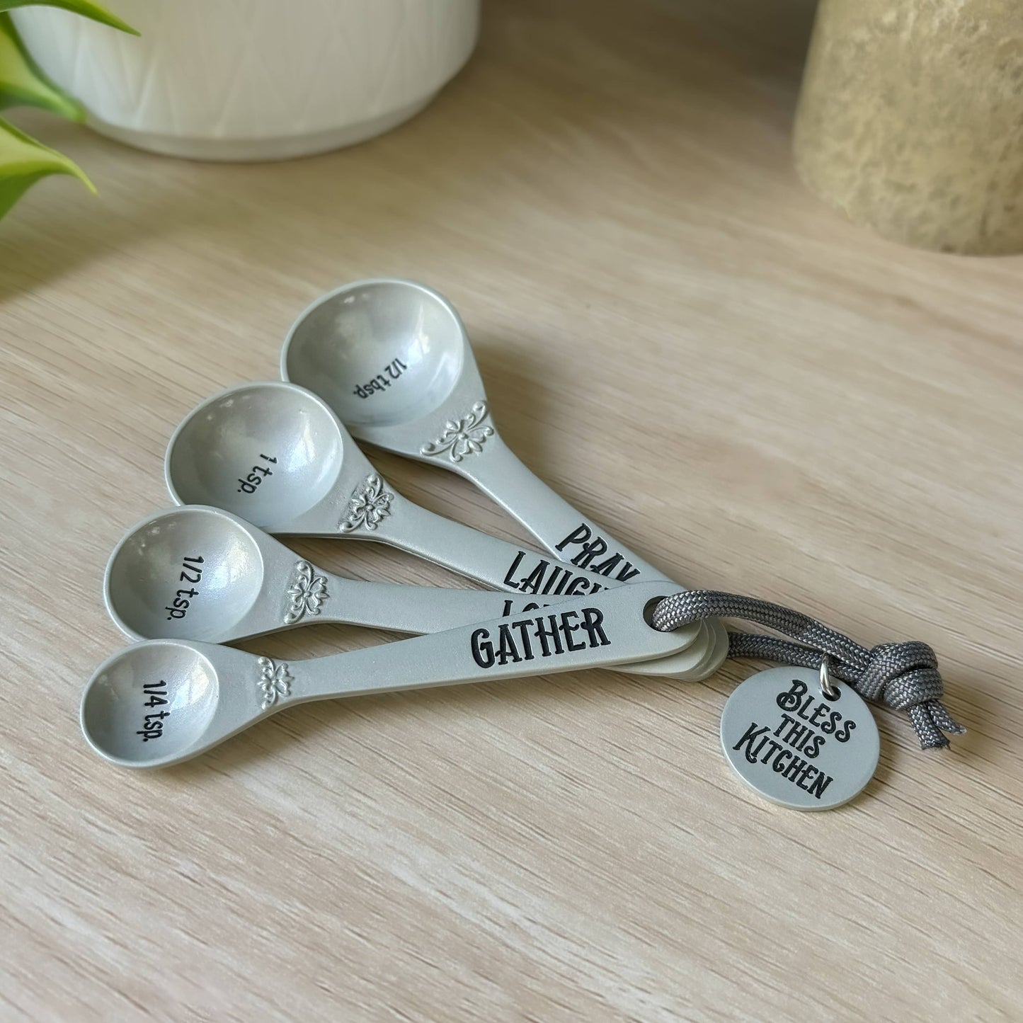 Set of five measuring spoons with engraved text on a wooden surface, accompanied by a tag that reads 'Bless this Kitchen'.