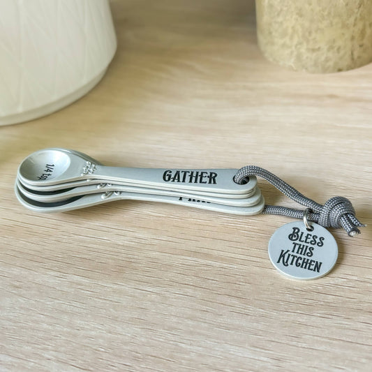 Set of measuring spoons with 'Gather' and 'Bless this Kitchen' charms on a wooden surface