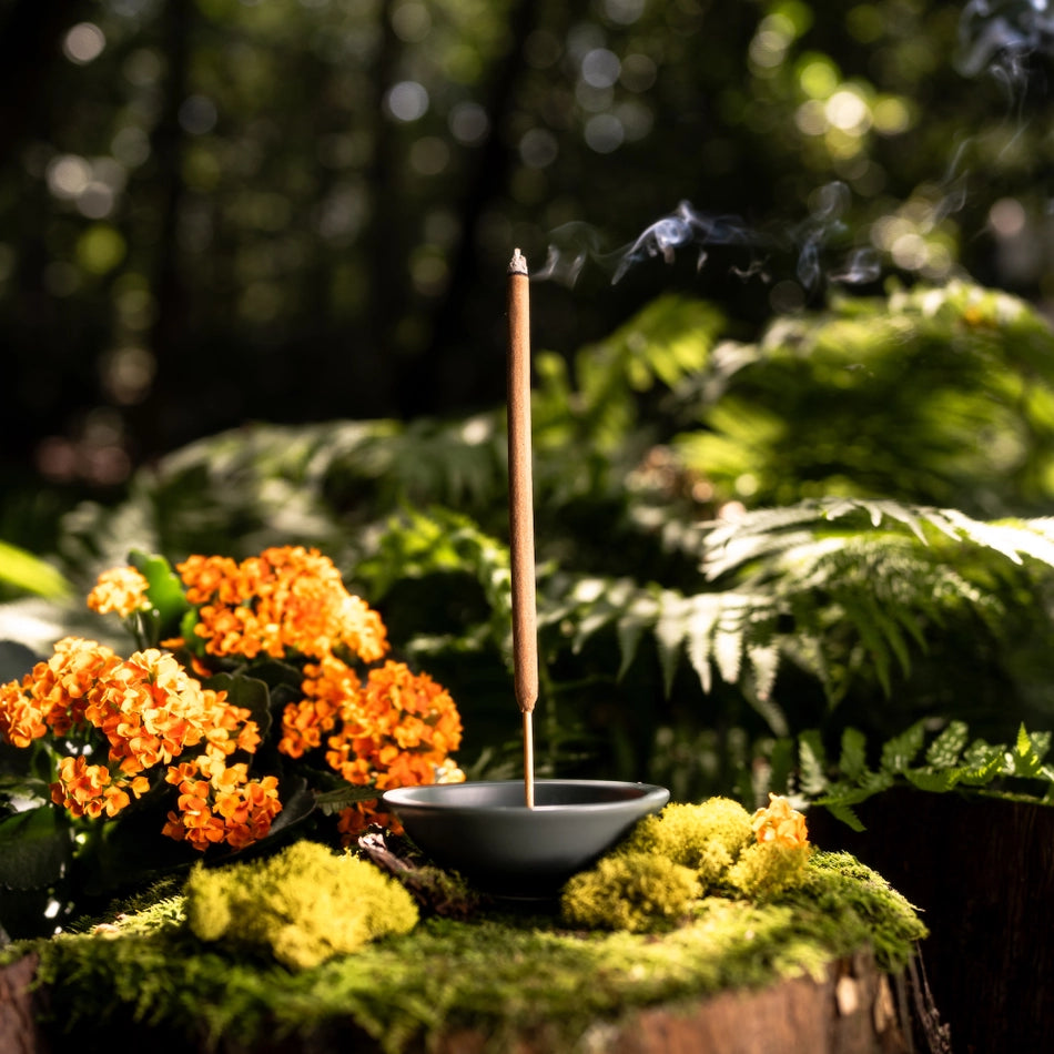 Incense stick burning on a mossy surface with flowers and greenery in the background