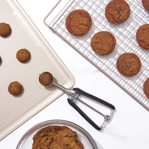 Baked ginger cookies on a cooling rack, cookie scoop, cookie sheet, and bowl of ground ginger.