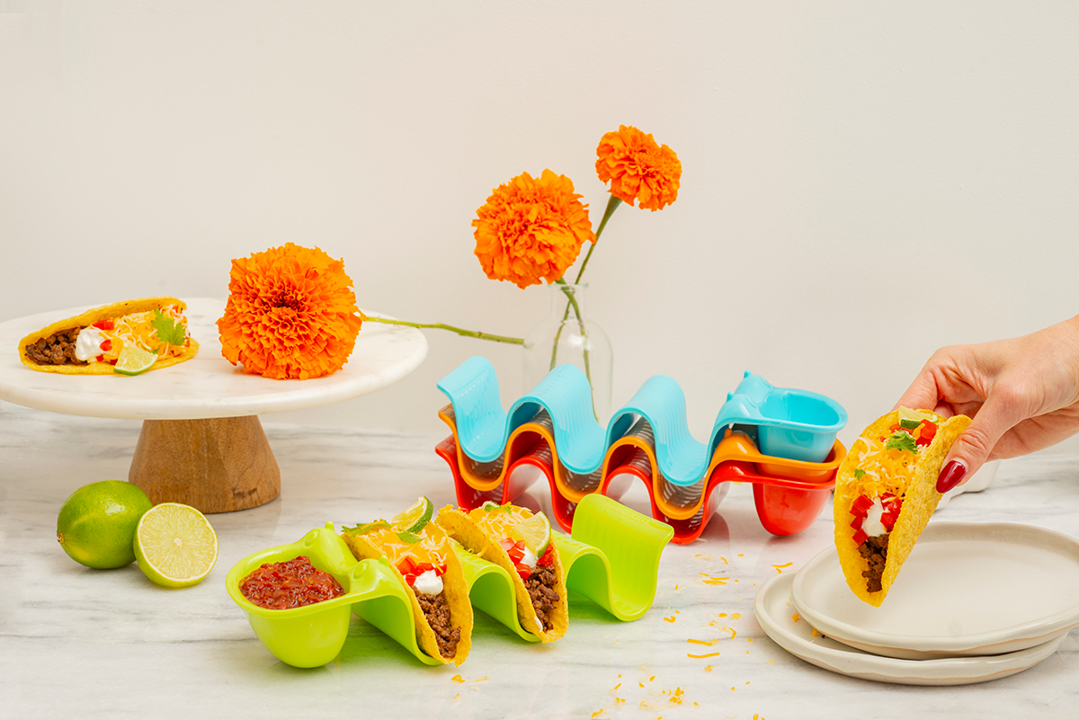Colorful plastic taco holders with mini tacos on a table with flowers and limes.