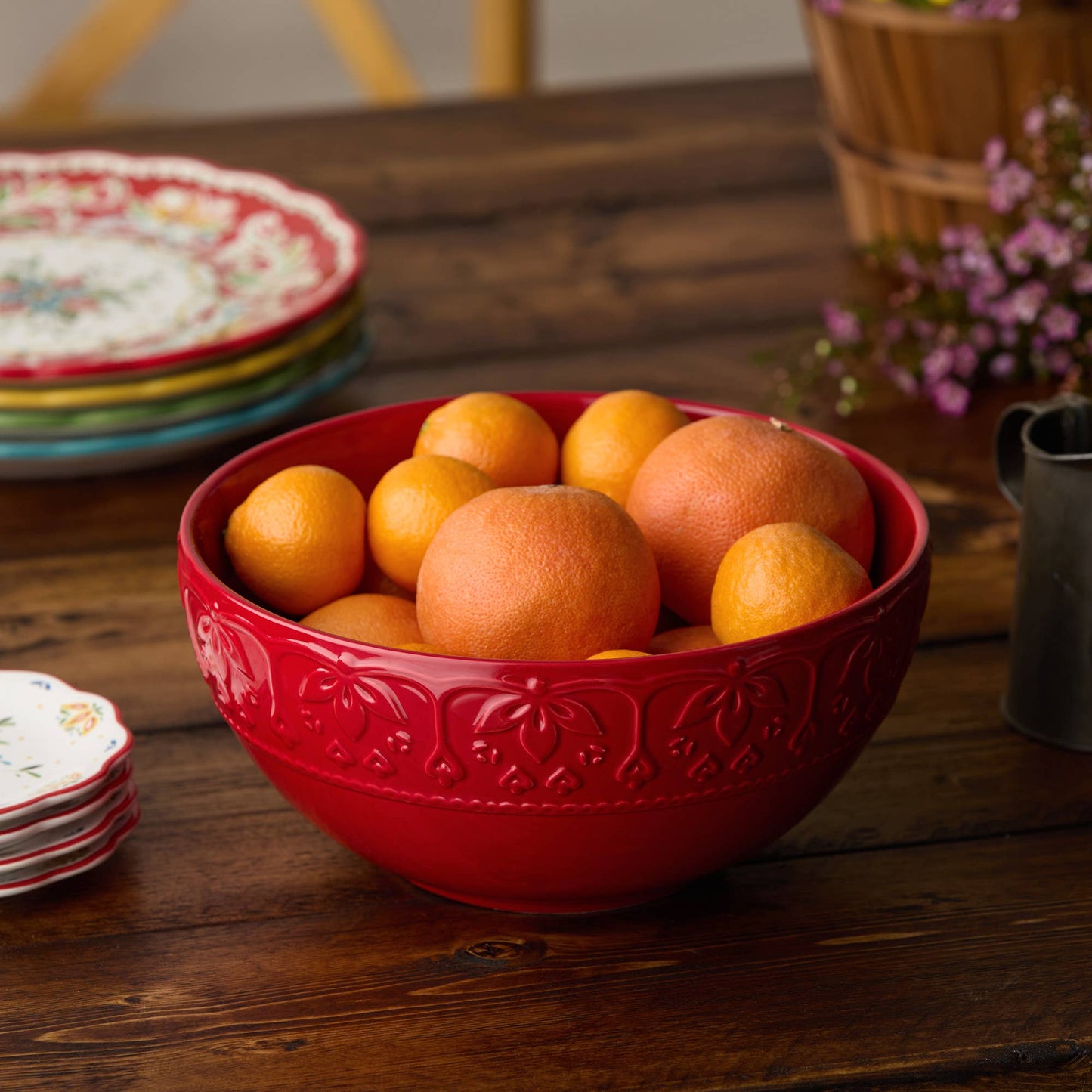 Red bowl filled with oranges on a wooden table with decorative plates and flowers in the background.