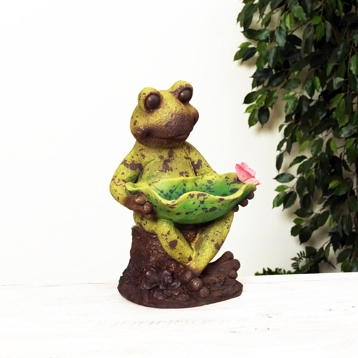 Frog statue holding a pink flower with green leaves in the background