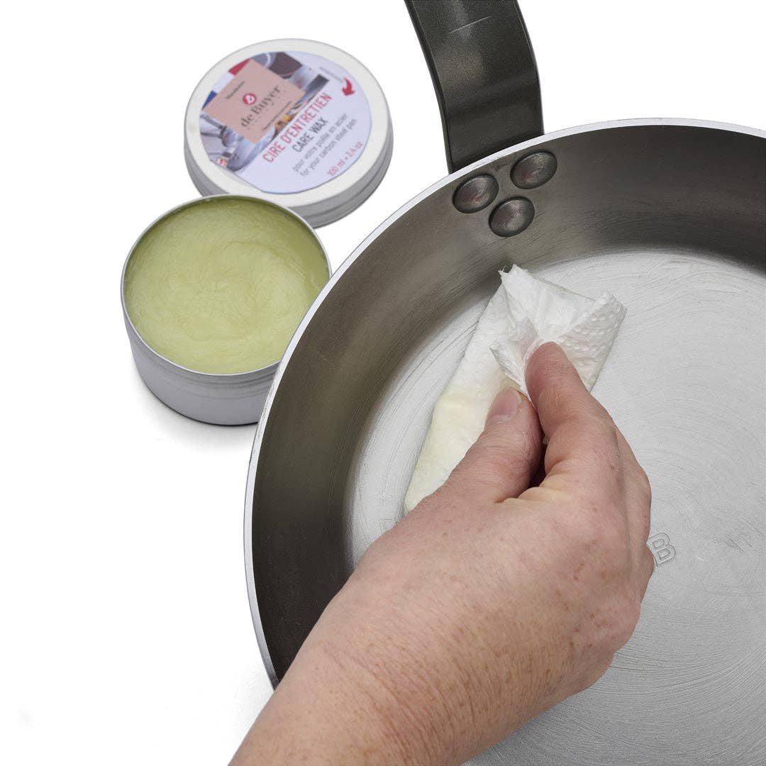 Person cleaning a frying pan with a cloth, next to a container of seasoning wax
