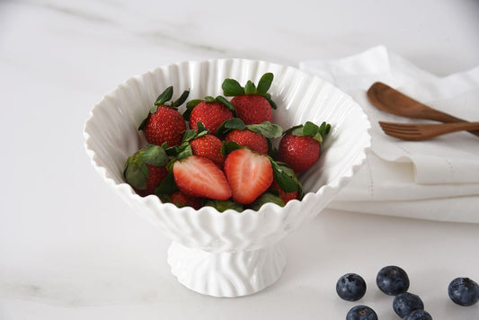 White bowl filled with strawberries on a white surface with blueberries and wooden utensils in the background.