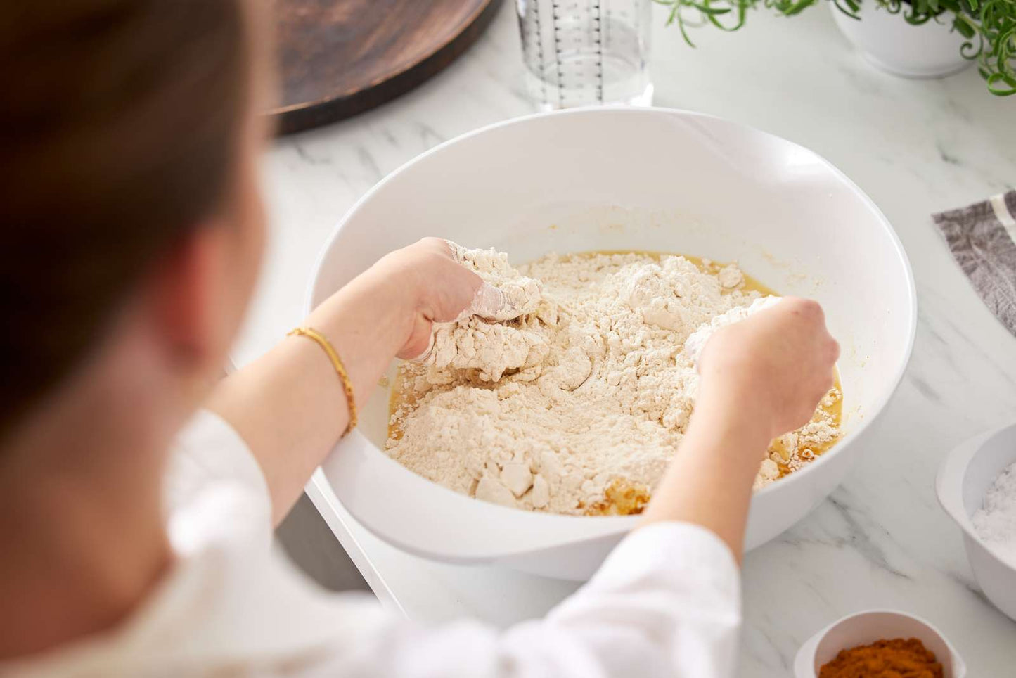 A person mixing dry ingredients, likely flour, into a wet batter in a large white bowl on a white marble countertop.