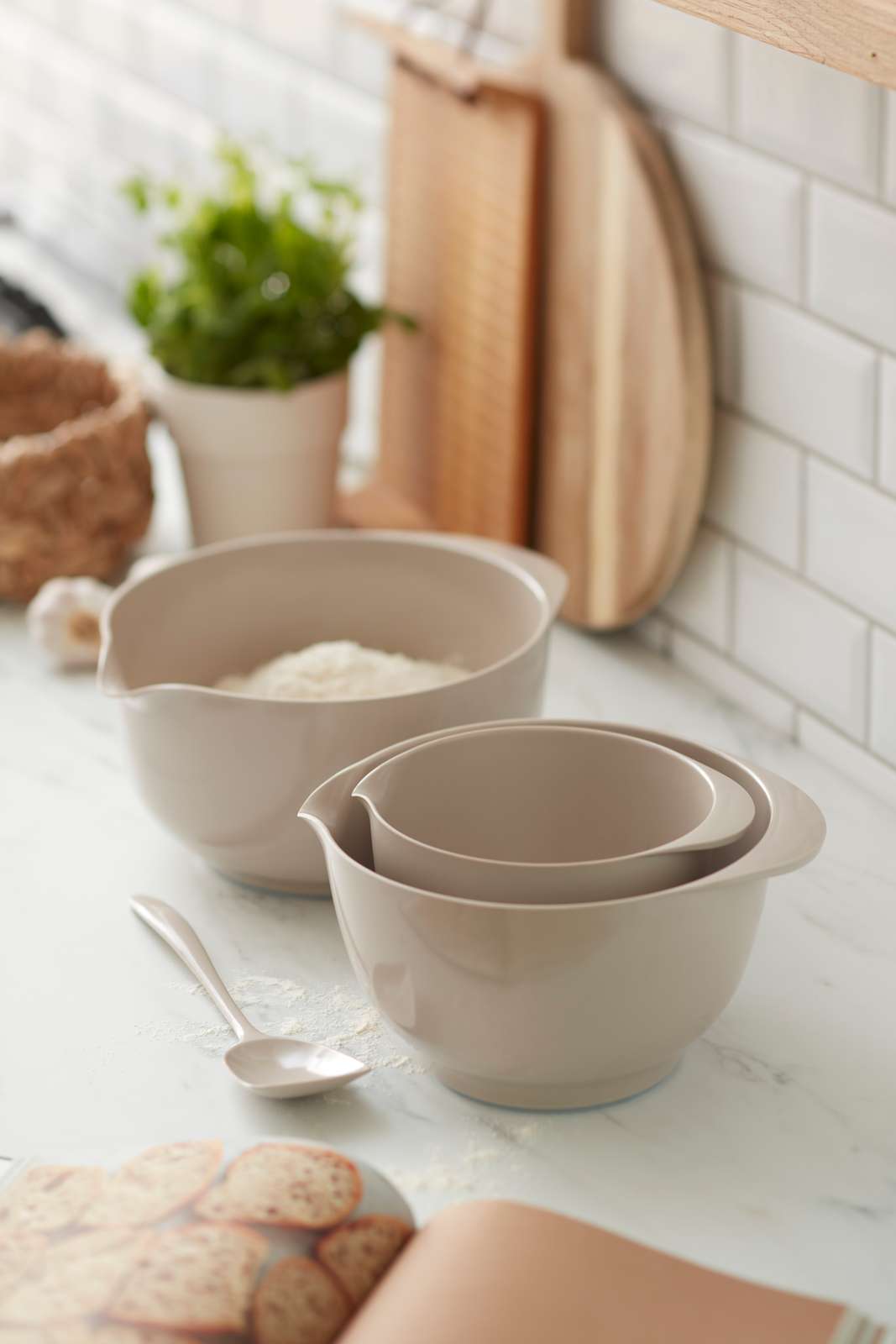 Three nested Rositi Margrethe Humus mixing bowls on a white marble kitchen counter, with a spoon, flour, cutting boards, and a small plant in the background.