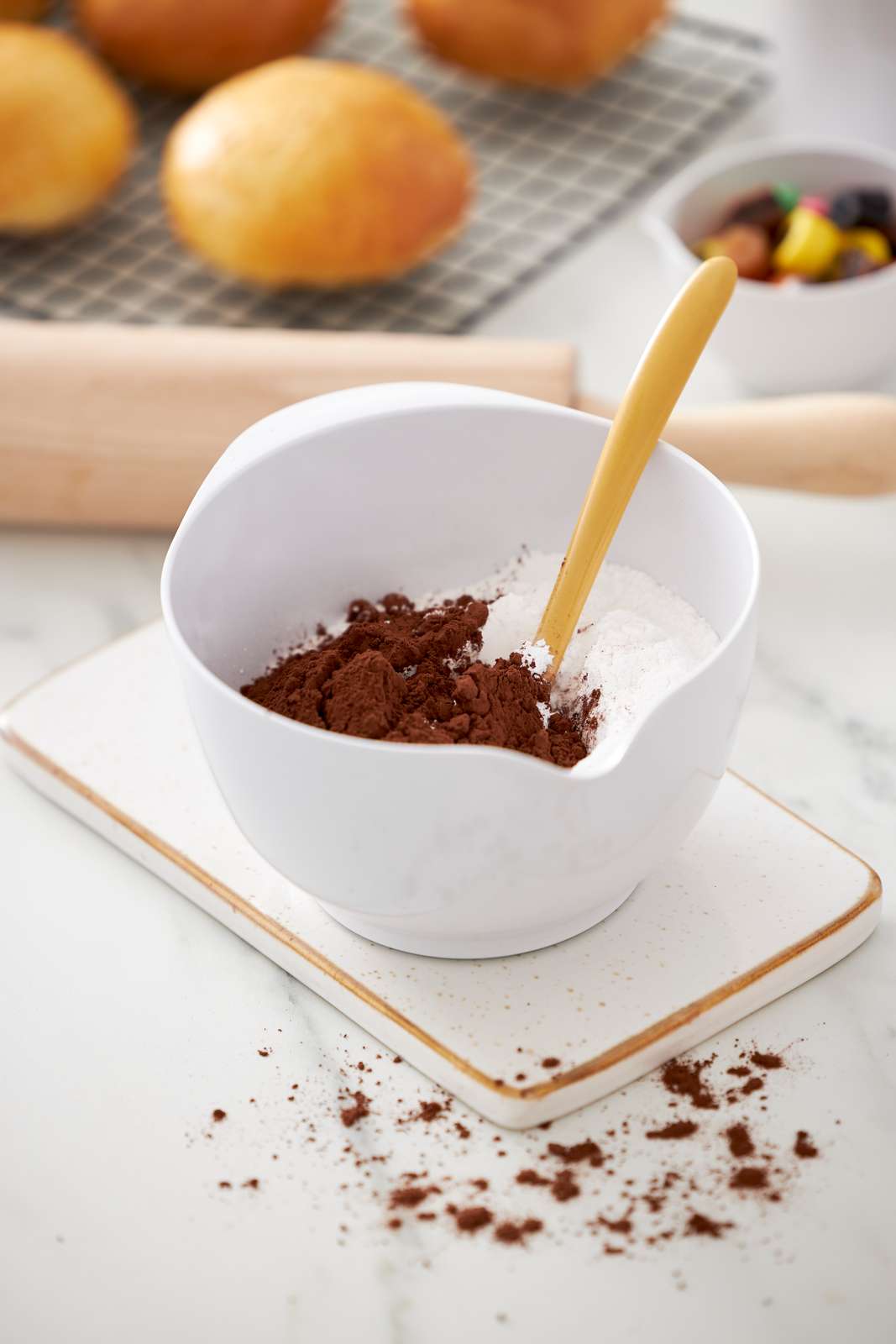 A white Rosti Margrethe bowl containing cocoa powder and flour with a yellow spatula, set on a square white coaster on a marble countertop. In the blurred background, golden-brown baked rolls sit on a cooling rack.