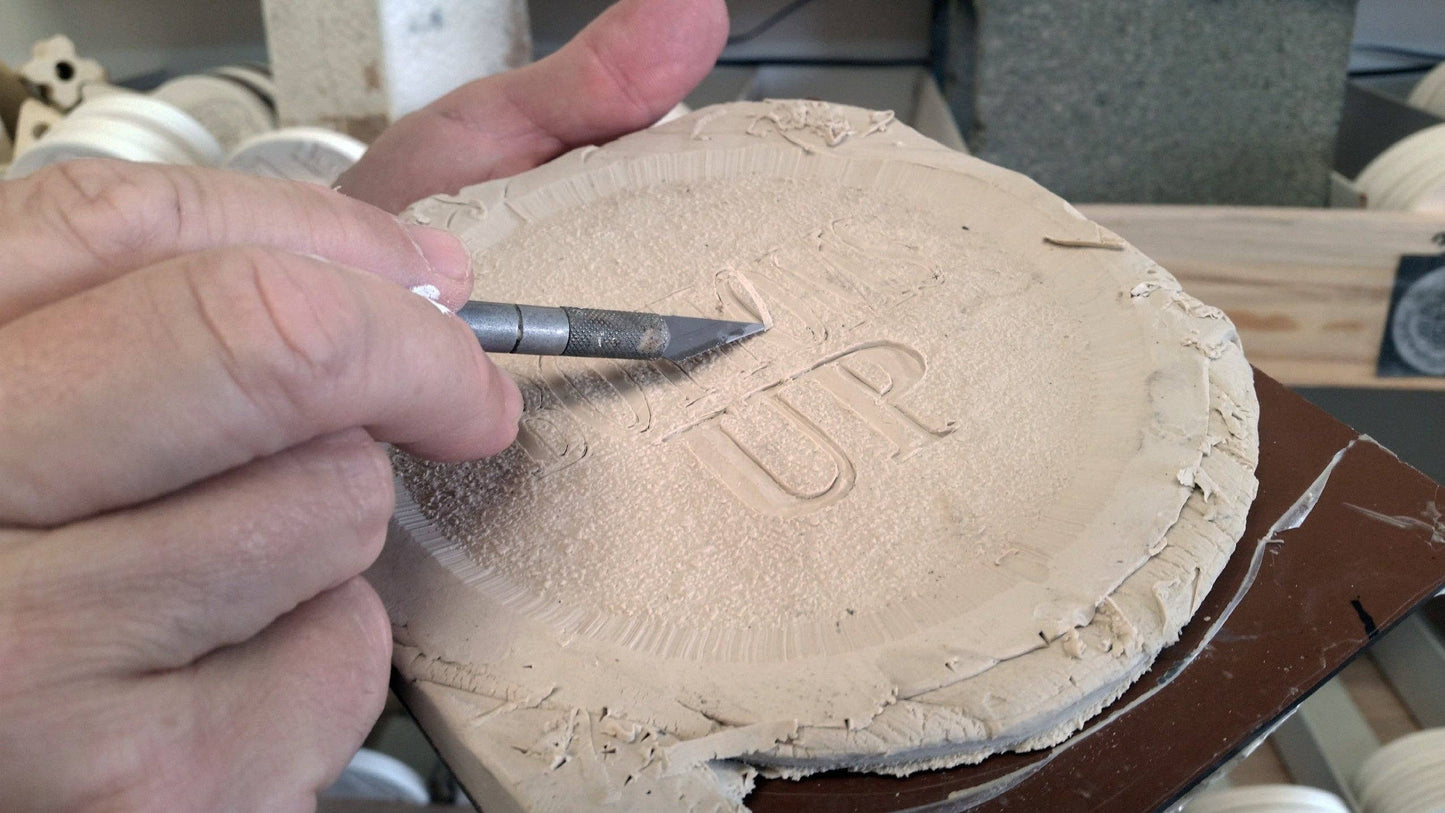 Person carving into a piece of clay with a tool, showing the word 'UP' being engraved.