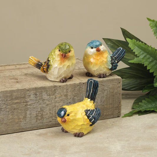 Three colorful bird figurines on a wooden surface with green leaves in the background.