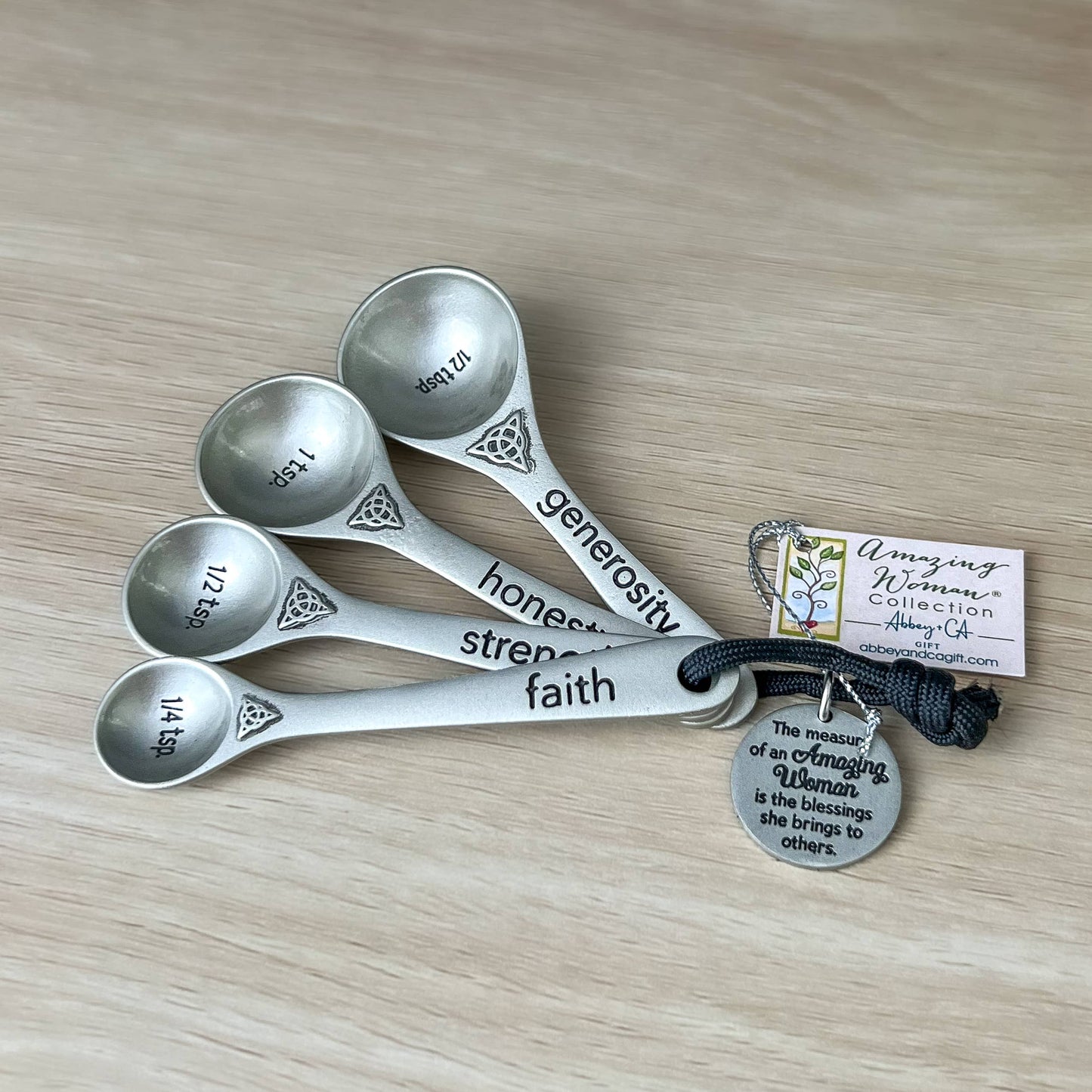 Set of silver measuring spoons with engraved words on a wooden surface, accompanied by a tag.
