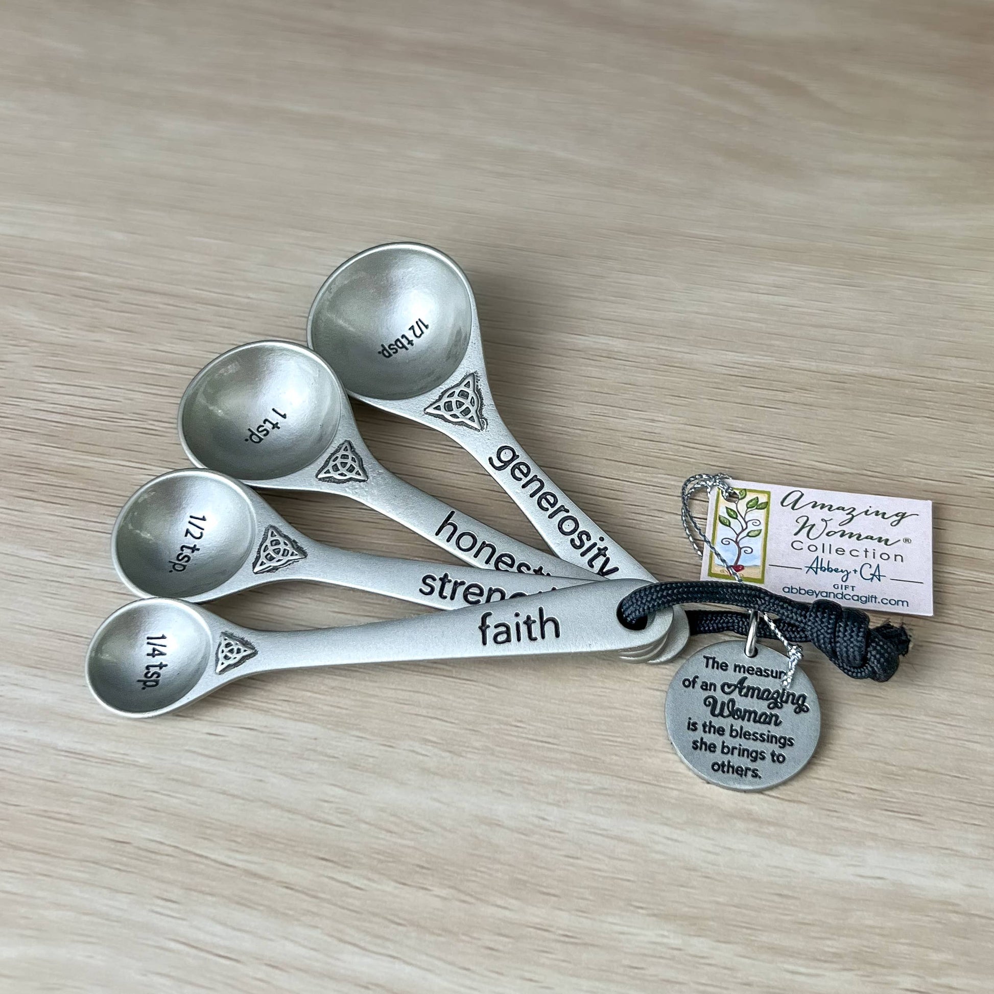 Set of silver measuring spoons with engraved words on a wooden surface, accompanied by a tag.