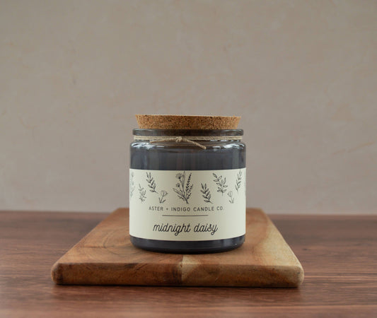 Candle jar labeled 'midnight daisy' on a wooden board with a beige background
