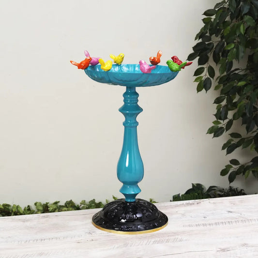 Turquoise bird bath with colorful bird figurines on a white surface with greenery in the background