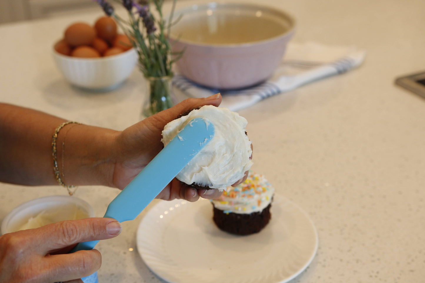 Person frosting a cupcake with a blue spatula on a kitchen counter.