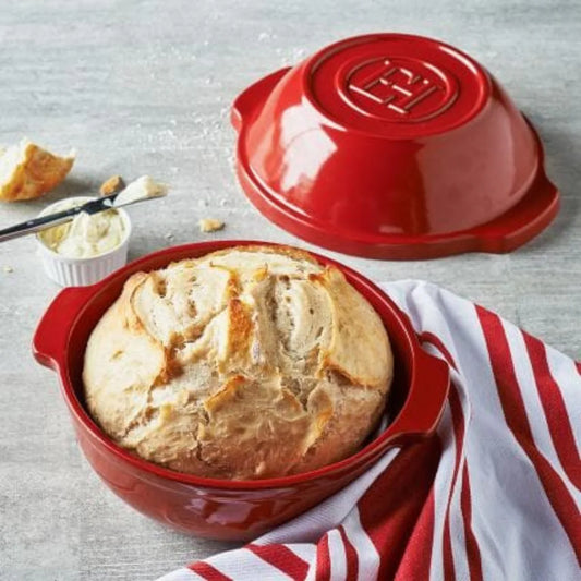 Red ceramic bread bowl with a loaf of bread inside, on a gray surface with a red and white striped towel.