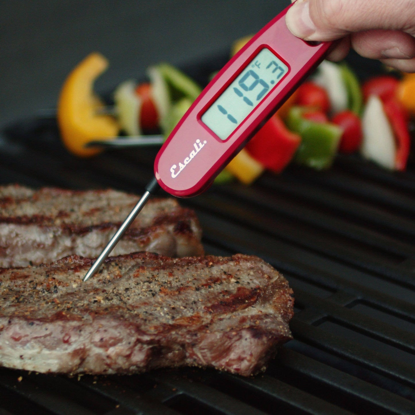 Digital thermometer reading 119.3°C being used to check the temperature of a steak on a grill with vegetables in the background.