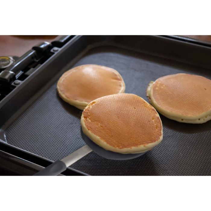 Three pancakes being cooked on a griddle with a spatula.
