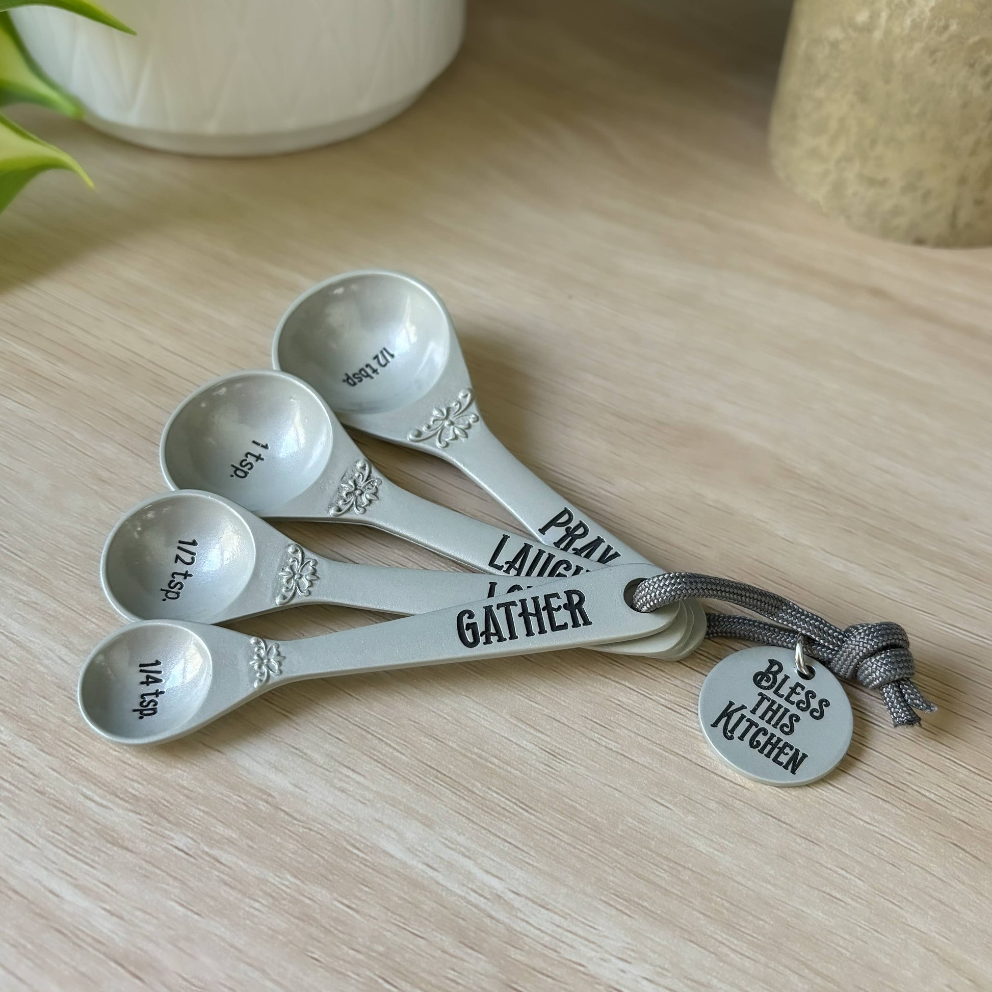 Set of five measuring spoons with engraved text on a wooden surface, accompanied by a tag that reads 'Bless this Kitchen'.