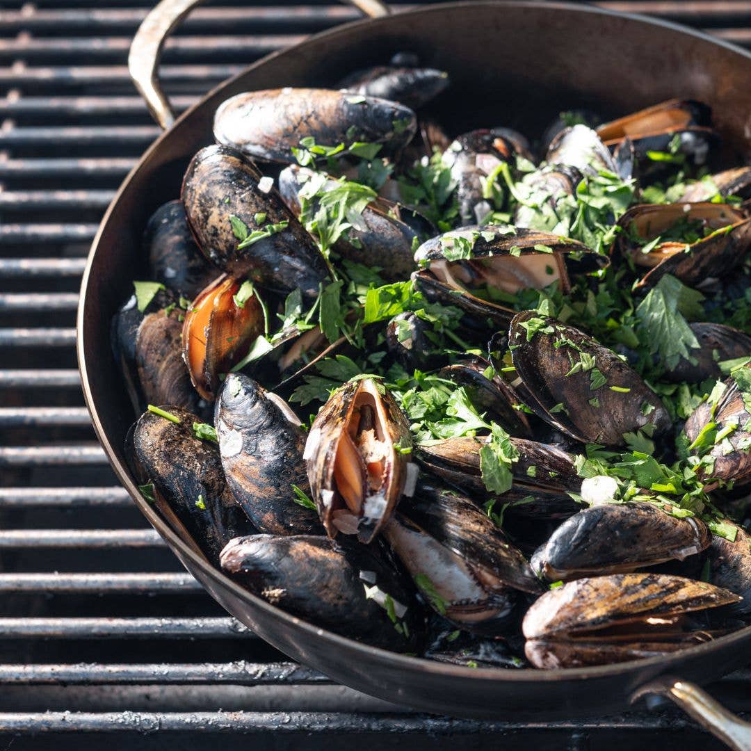 Mussels cooked in a dark cast-iron skillet on a grill grate, topped with chopped fresh green parsley.