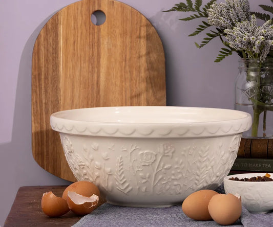 White embossed bowl on a wooden surface with eggs and a cutting board in the background.