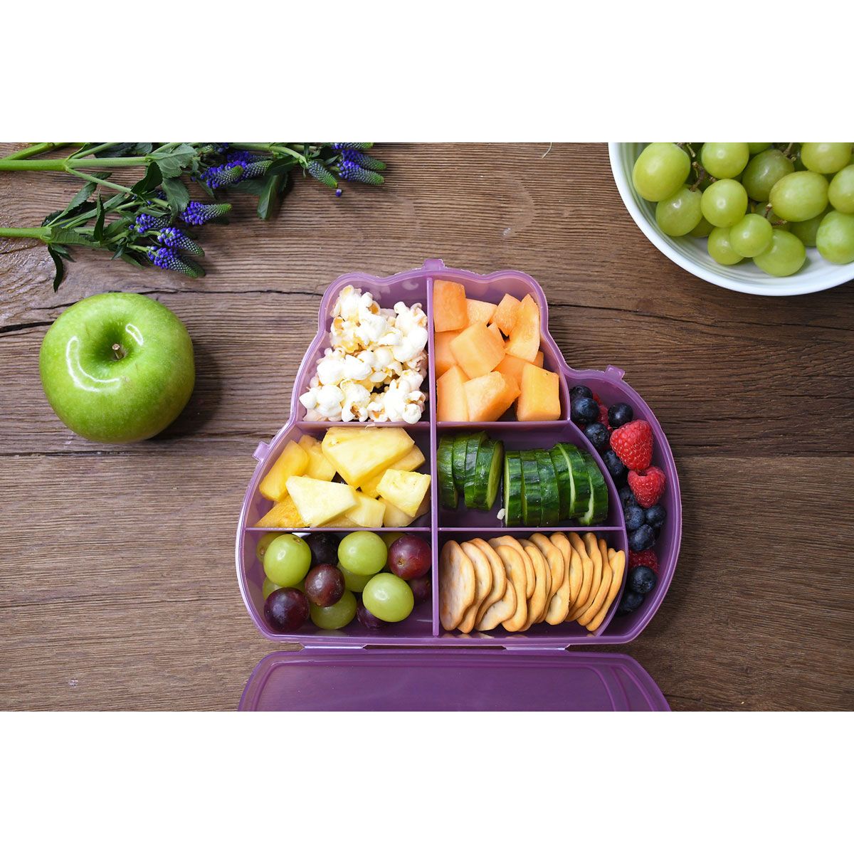 Purple cat shaped snack box filled with various fruits and snacks on a wooden table.