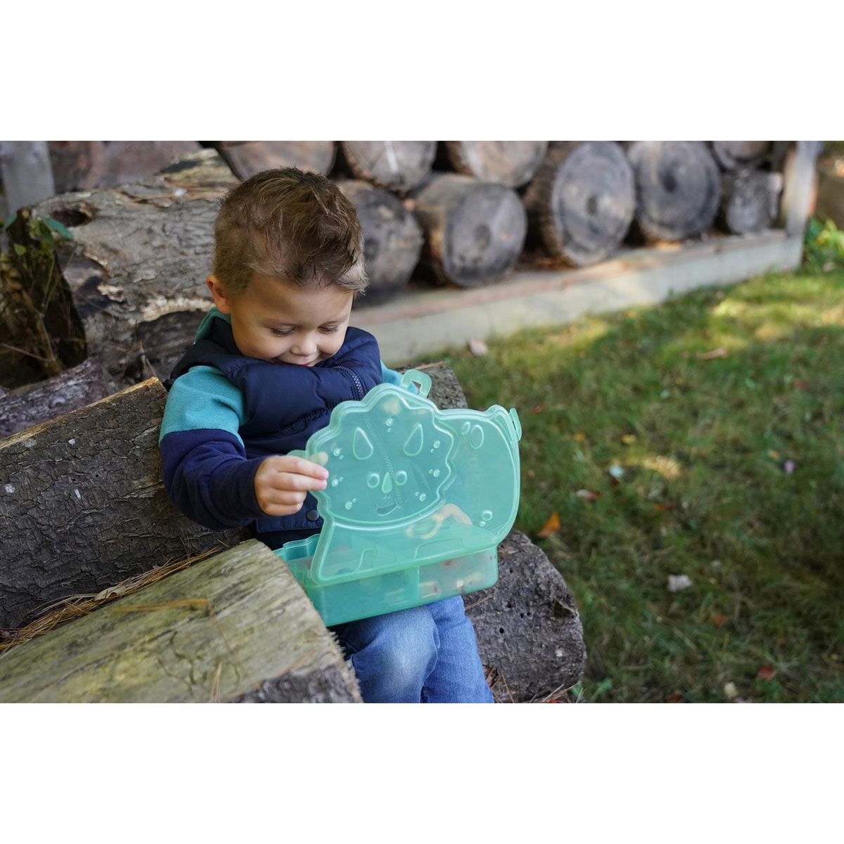 Child holding a Melii Dino Snackle Box in an outdoor setting with logs and grass.