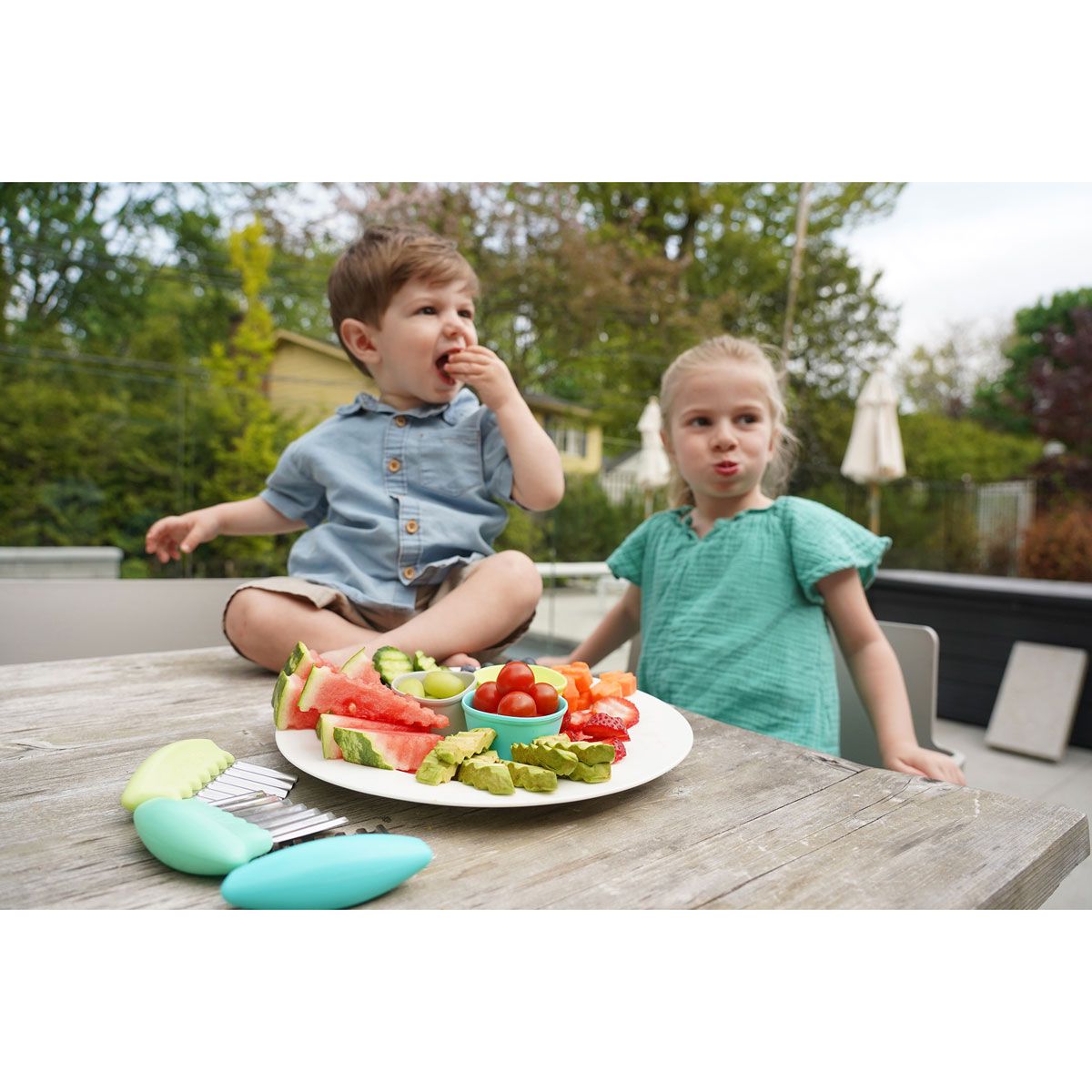 Two children eating a plate of fruit and vegetables outdoors.
