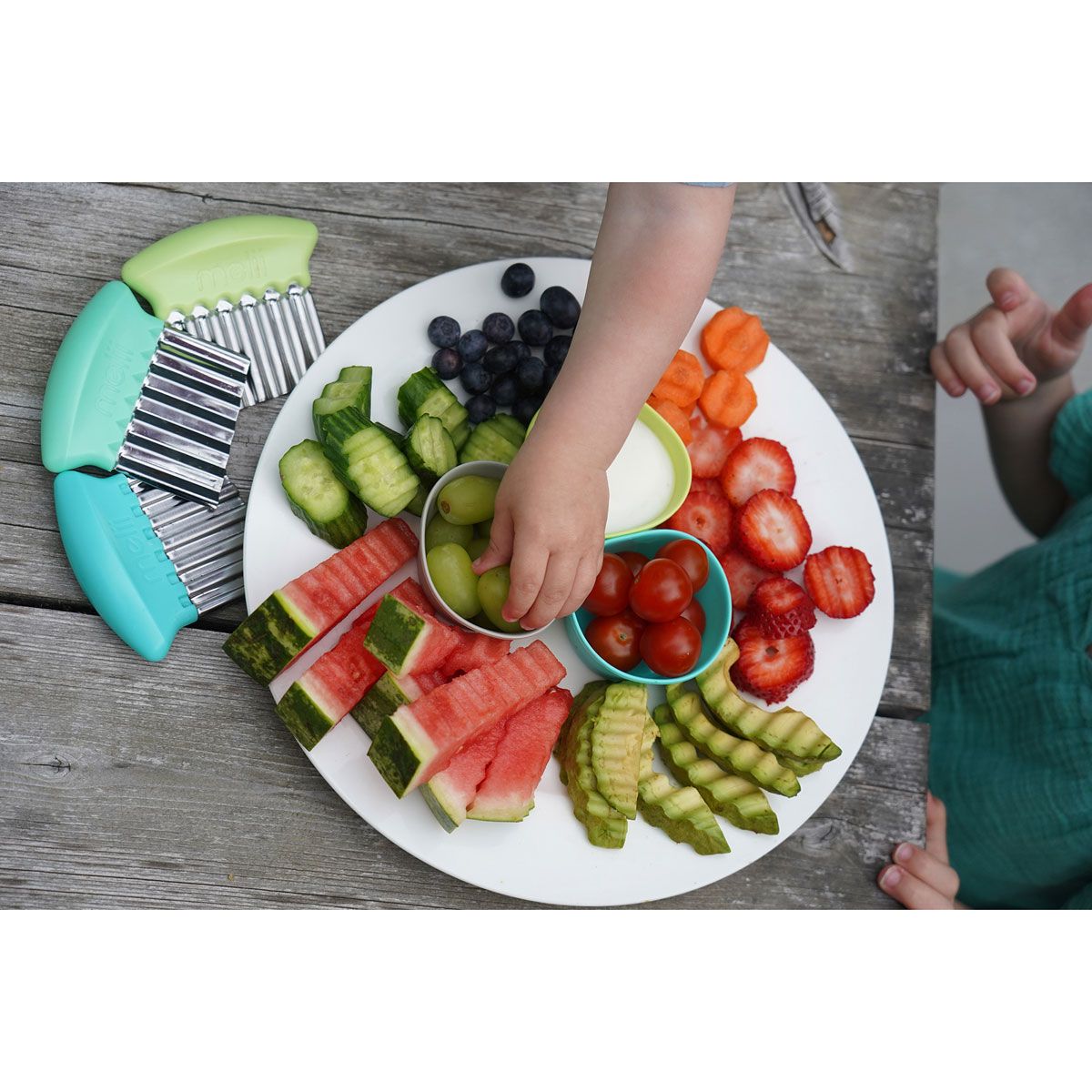 Assorted fruits and vegetables on a plate with children's hands reaching for them.