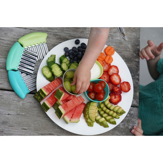 Assorted fruits and vegetables on a plate with children's hands reaching for them.