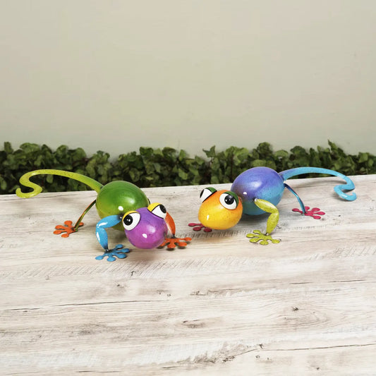 Colorful metal gecko figurines on a wooden surface with a green foliage background
