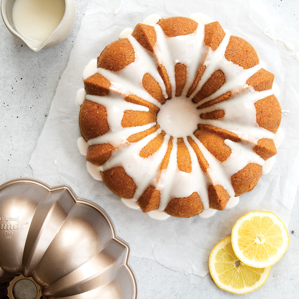 Bundt cake with white icing on a light gray surface with lemon slices and a cake pan.