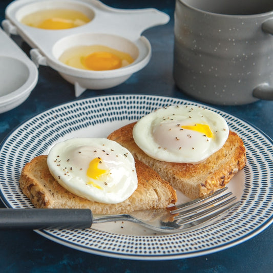 Two poached eggs on toast with a fork on a plate, next to a poaching pan and mug.