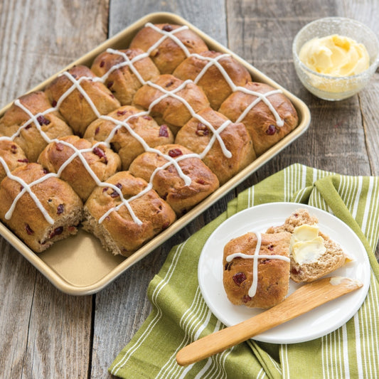 Hot cross buns on a baking tray with one cut open on a plate, accompanied by a dish of butter.