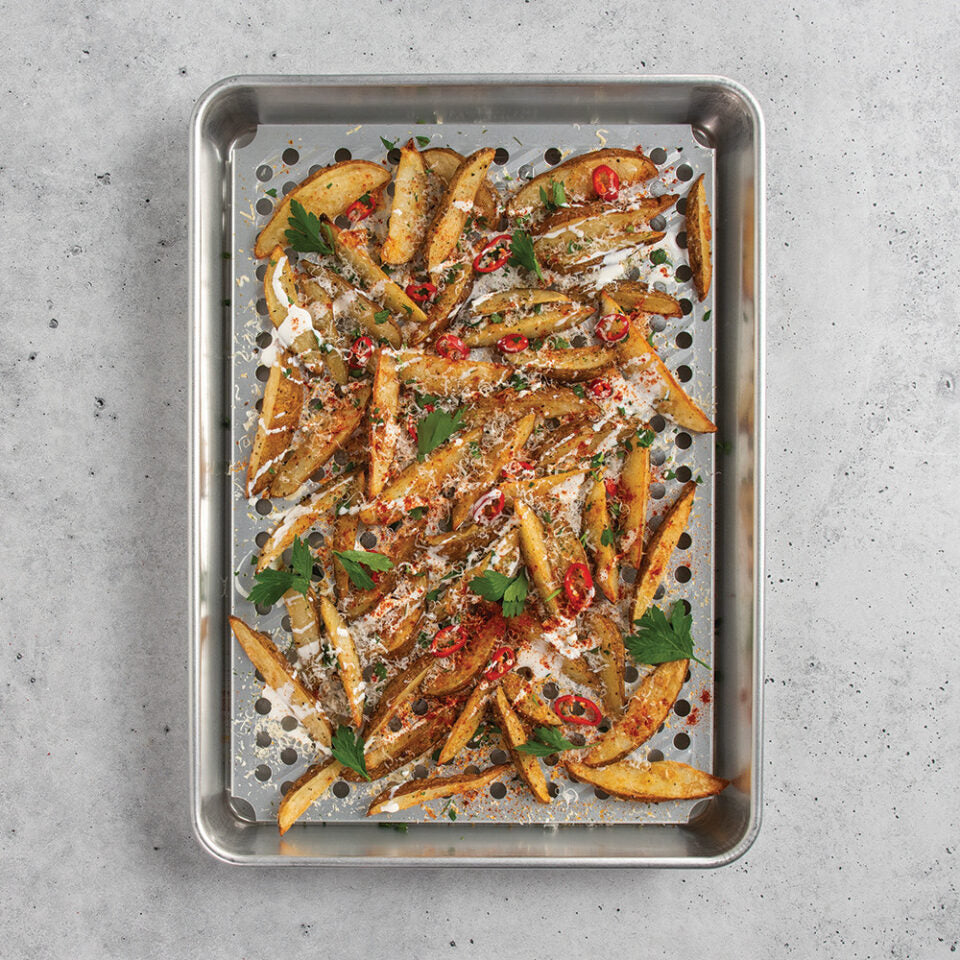 Metal baking sheet with roasted potato wedges garnished with herbs and red peppers on a gray surface.