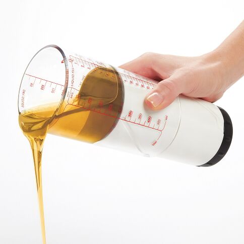 Hand pouring oil from a measuring jug with measurement markings on a white background