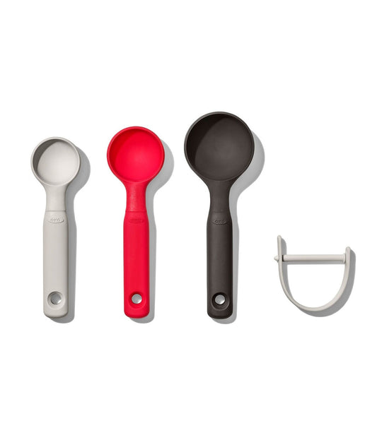 Set of four kitchen utensils including a white measuring spoon, red ladle, black ladle, and a white peeler on a white background.