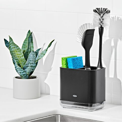 Black caddy with cleaning utensils on a kitchen counter next to a plant