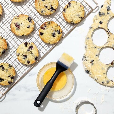Blueberry muffins on a cooling rack with a brush and bowl of butter on a marble surface.